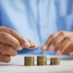 Hand placing a coin into a glass jar labeled investing representing micro asset growth